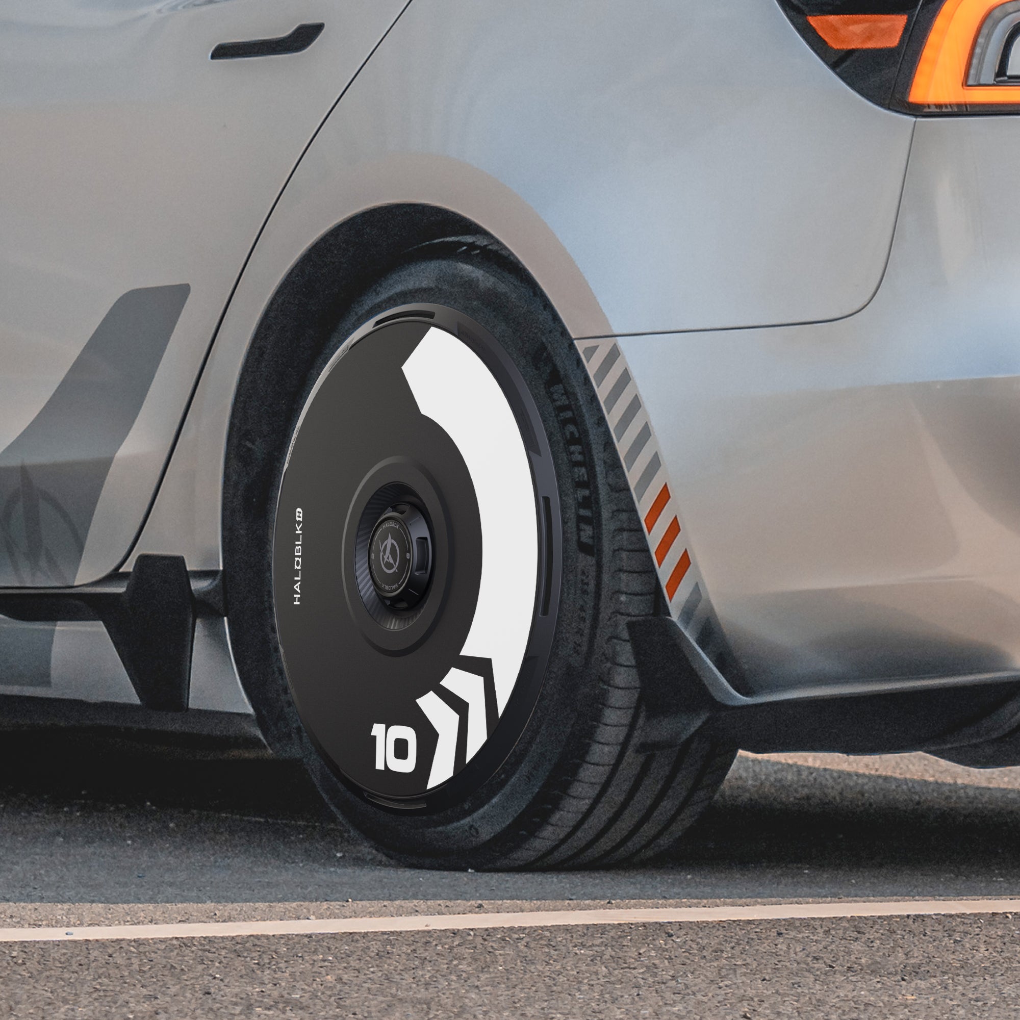 Close-up of HALODISC 2 Wheel Covers by HALOBLK Store on a Tesla Model 3 Highland 19" Nova Wheel, featuring an Arrow design with black and white geometric patterns and the number “10”, plus orange accents on the car’s side.