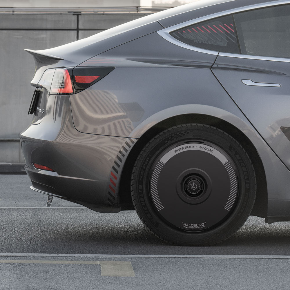 A close-up of a Tesla Model 3 (2019-2023) with HALODISC 2 Silver Track wheel covers by HALOBLK Store, featuring circular patterns and logo, showcases the 19" sport wheels against an urban backdrop.