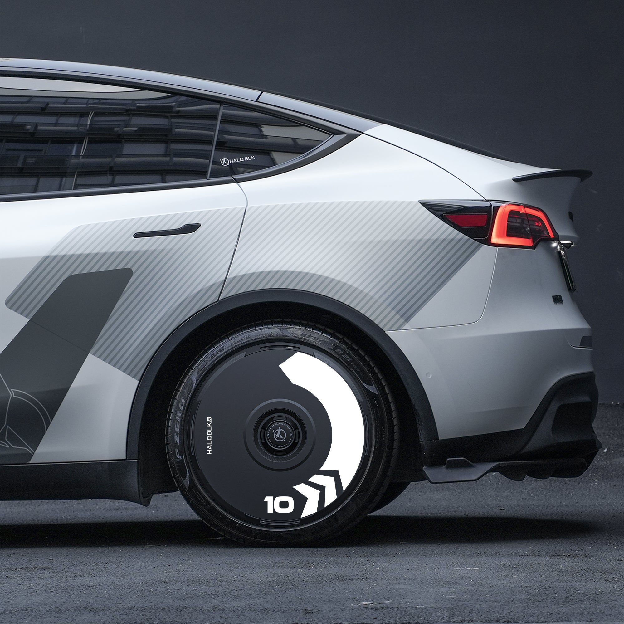 A close-up side view of a modern grey Tesla Model Y with HALODISC 2 Wheel Covers (Arrow design) from HALOBLK Store on 19" Gemini wheels, featuring a unique black and white pattern marked with "10" and arrows; rear taillight illuminated.