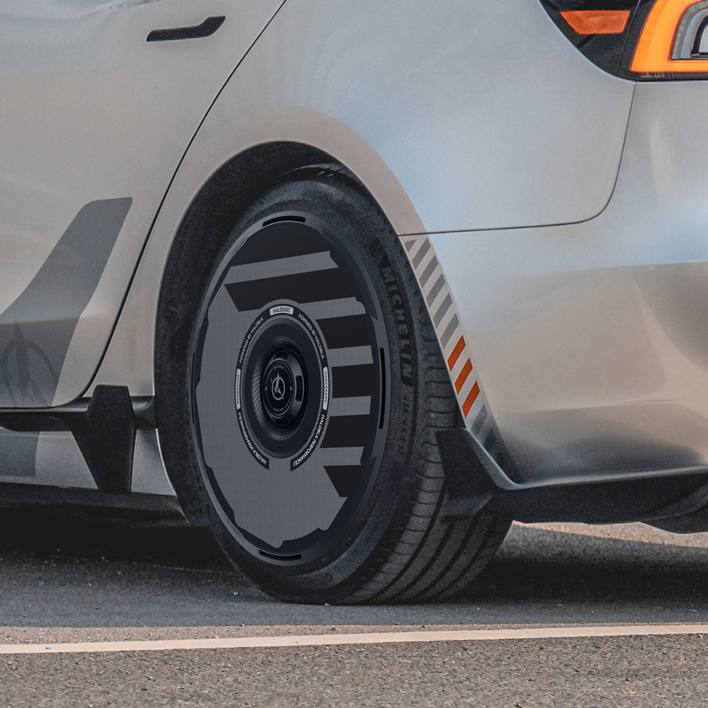Close-up of a silver Tesla Model 3 Highland with HALODISC 2 Wheel Covers (19" Nova) by HALOBLK Store, featuring black and gray geometric patterns and Michelin tires. Orange stripes accent the fender.