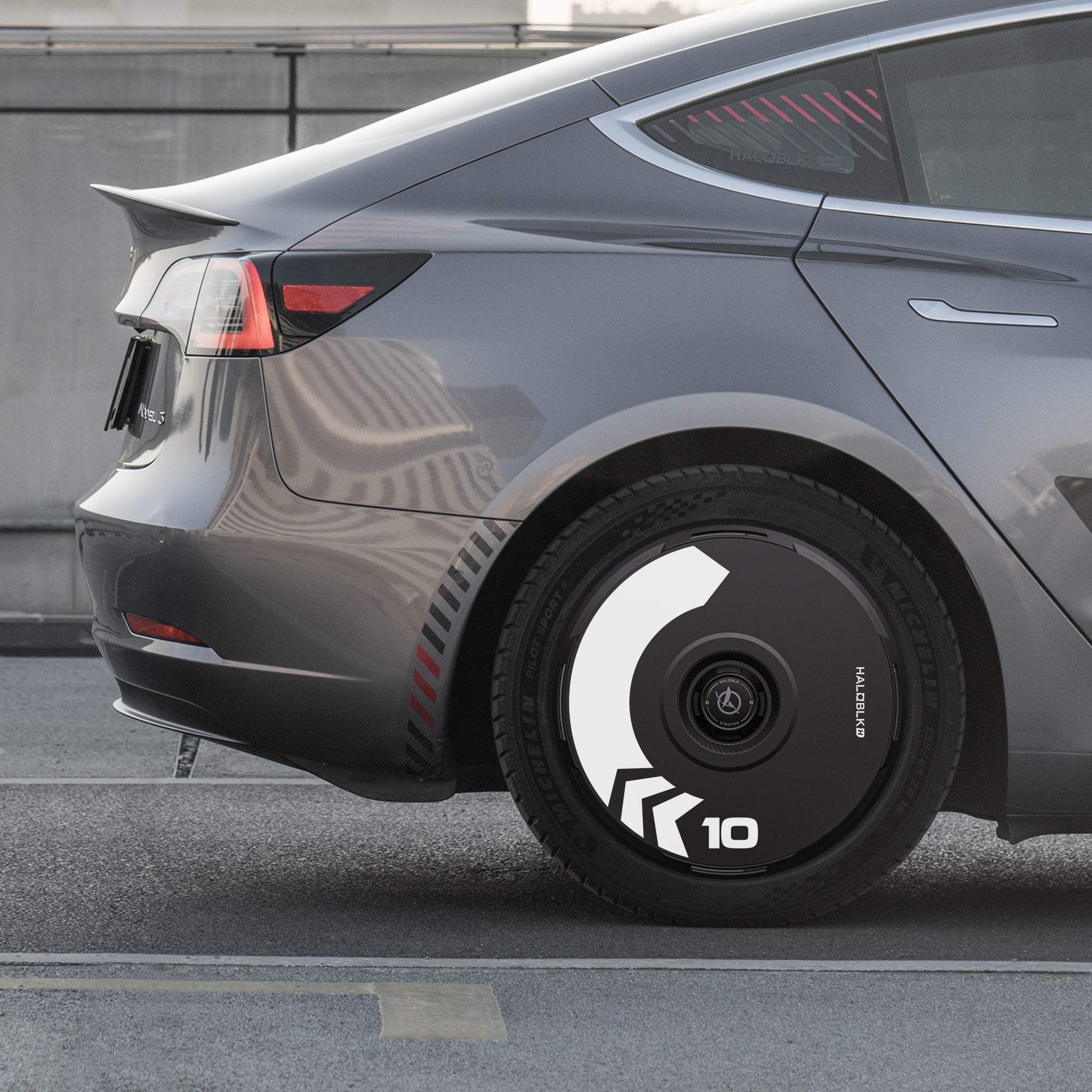 A gray Tesla Model 3 (2019–2023) with HALODISC 2 Arrow Wheel Covers by HALOBLK Store is parked in an urban concrete setting, showcasing bold black and white geometric patterns with the number 10 on the covers.