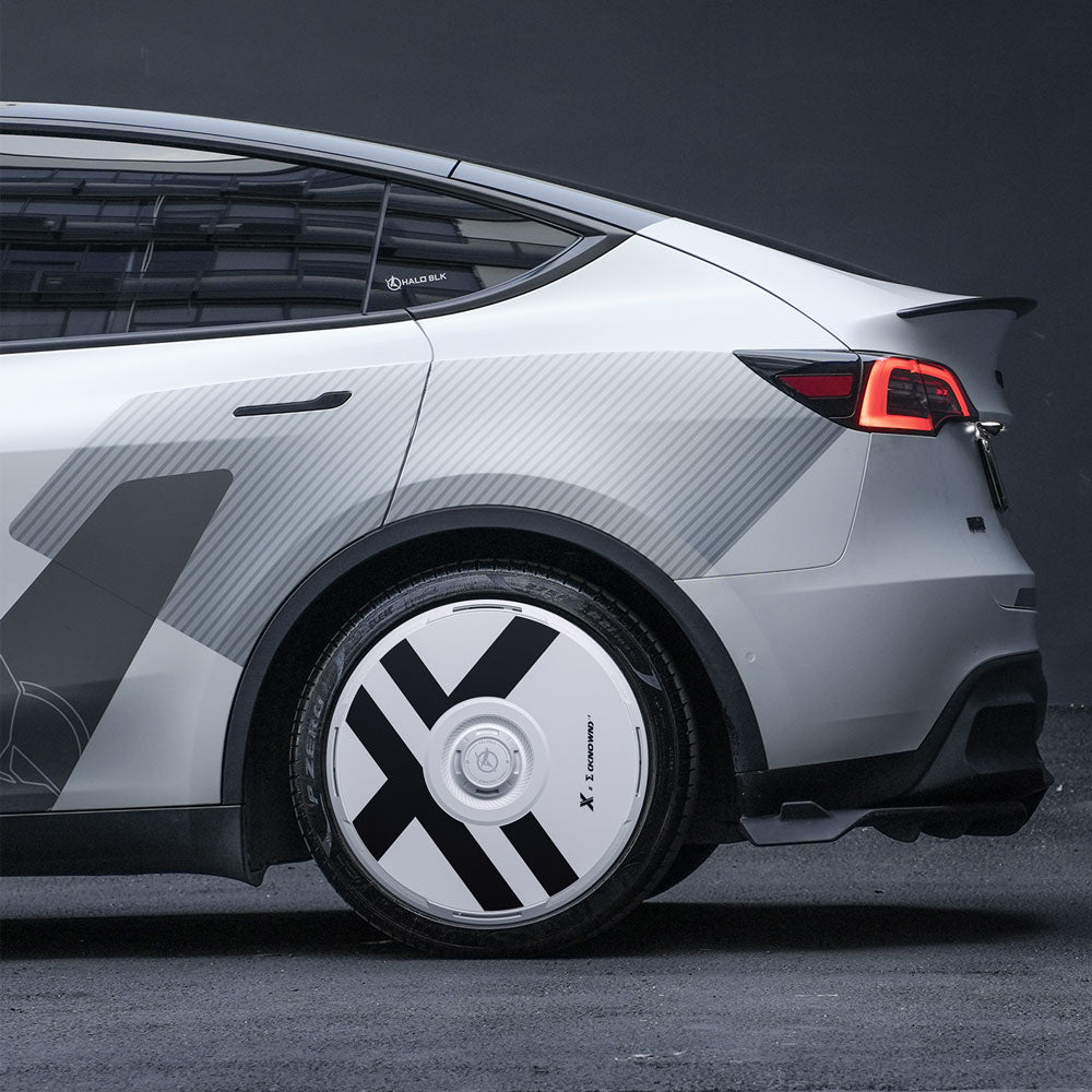 A side view of a silver Tesla Model Y with HALODISC 2 Wheel Covers for 21" Überturbine Wheels from HALOBLK Store, featuring bold black and white geometric X-Factor patterns. The car has sleek lines and dark-tinted windows.