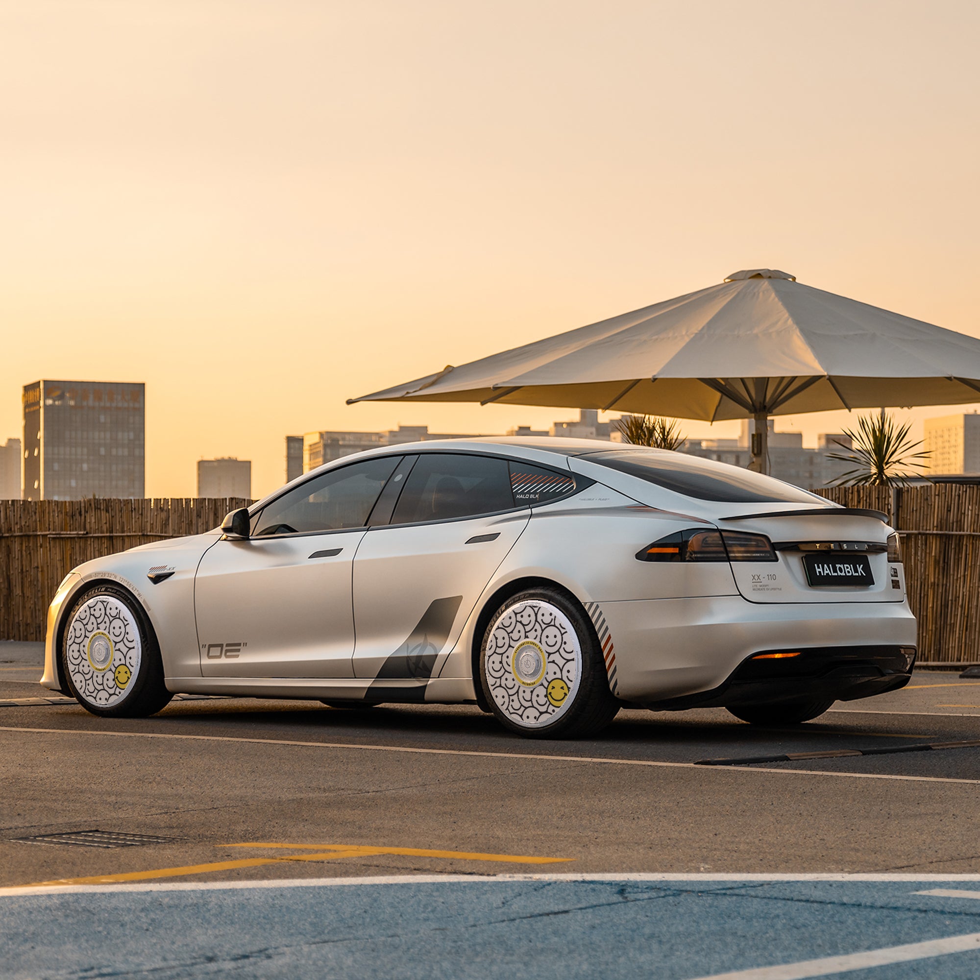 A sleek silver Tesla Model S with HALODISC 2 Wheel Covers by HALOBLK Store is parked at sunset in an urban setting, city buildings, a large white umbrella, and a bamboo fence in the background.