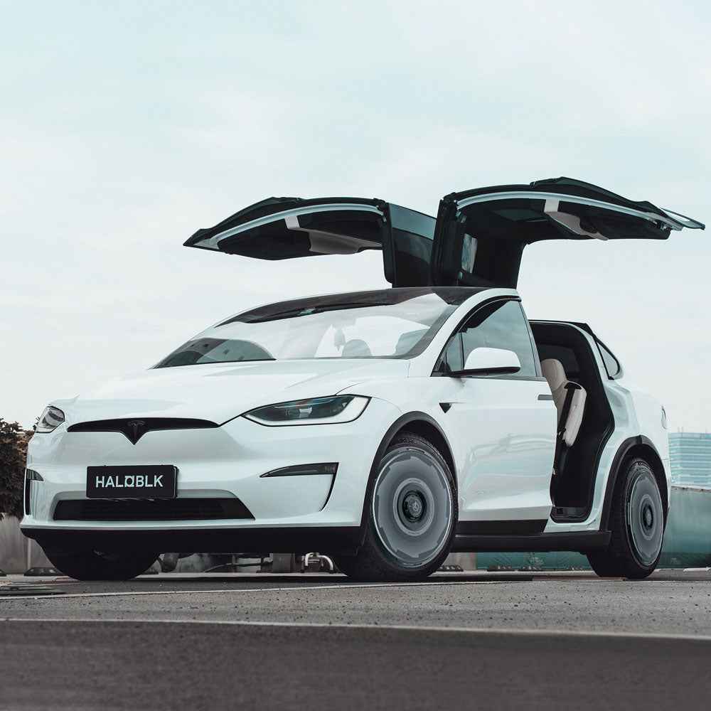 A white Tesla Model X (2021-2025) with 20" HALODISC 2 Vision-GT wheel covers from HALOBLK Store and a HALOBLK license plate, parked with falcon-wing doors open under a cloudy sky. Front and side are visible.