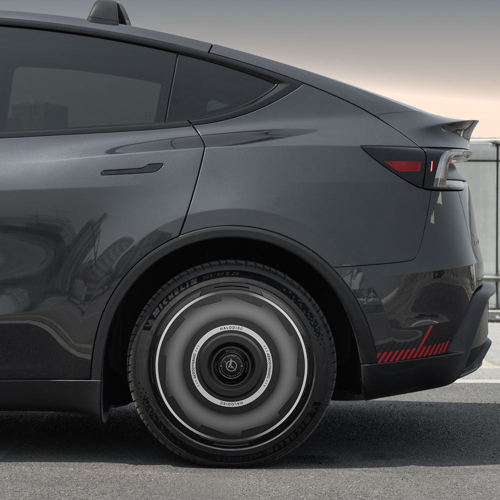 A close-up of the rear side of a Tesla Model Y with HALODISC 2 Wheel Covers for Juniper 19" Crossflow Wheels by HALOBLK Store, showcasing the futuristic circular pattern design. The vehicle is parked on asphalt with a blurred urban background.