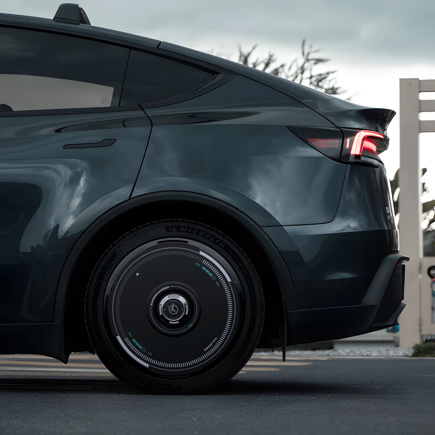 Close-up side view of a Tesla equipped with HALODISC 2 Wheel Covers from HALOBLK Store, highlighting the rear wheel and modern taillight, parked outdoors under an overcast sky with trees and a fence in the background.