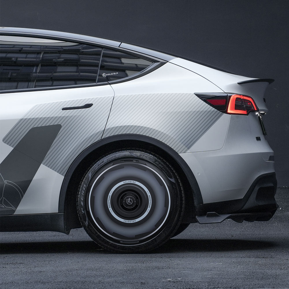 A close-up of the rear of a white Tesla Model Y with HALODISC 2 Wheel Covers by HALOBLK Store for 21" Überturbine Wheels, featuring a geometric decal, glowing tail lights, and a futuristic design, parked on a dark surface.
