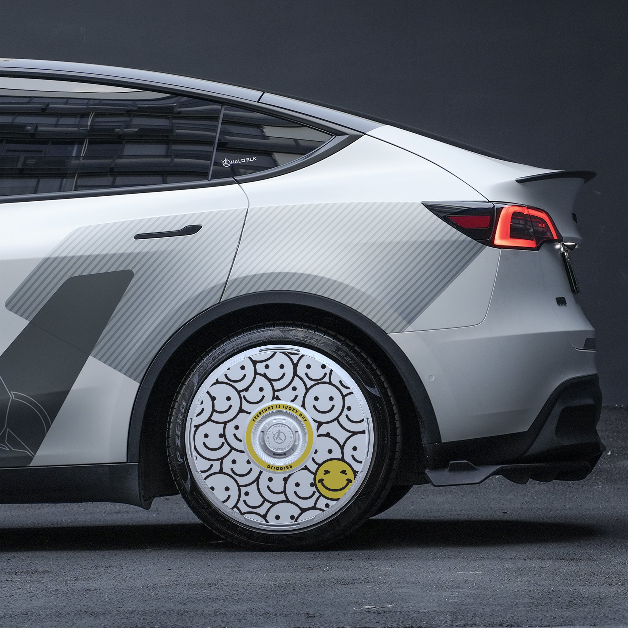 A white Tesla Model Y with HALODISC 2 Wheel Covers ("Lucky Day" by HALOBLK Store) featuring a grey geometric side design and a rear wheel cover patterned with black smiley faces and a standout large yellow smiley face, taillight illuminated in a dark setting.