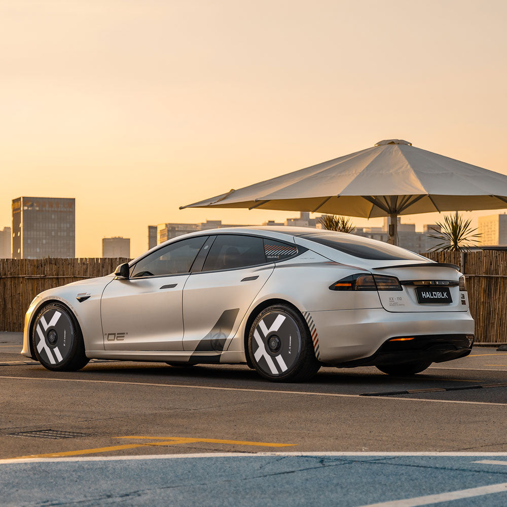 A silver Tesla Model S with HALODISC 2 Wheel Covers by HALOBLK Store is parked at sunset in an urban setting, near a large outdoor umbrella and wooden fencing, with city buildings in the background.
