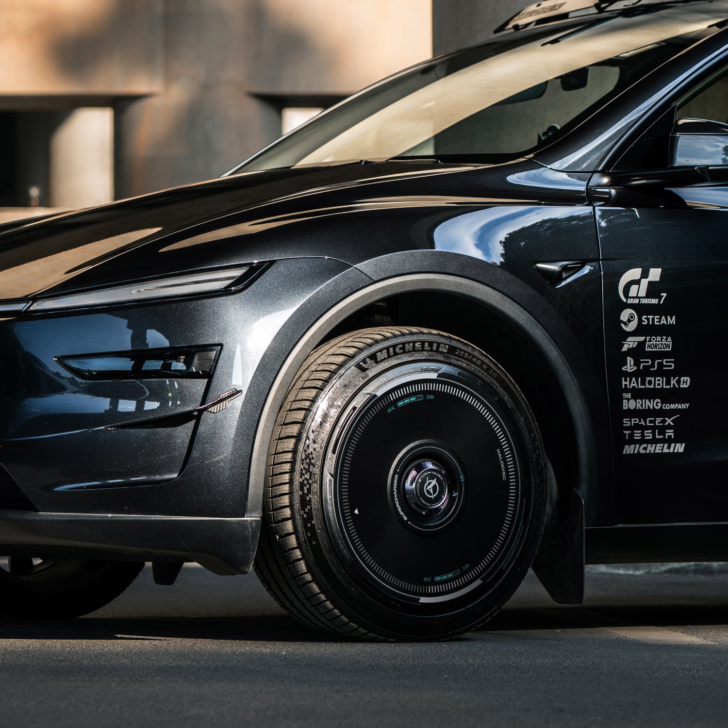 Close-up of a black car's front side, featuring HALODISC 2 Wheel Covers for Tesla by HALOBLK Store. Sponsor logos—Steam, PS5, SpaceX, and Tesla—appear on the door. The background is out of focus.