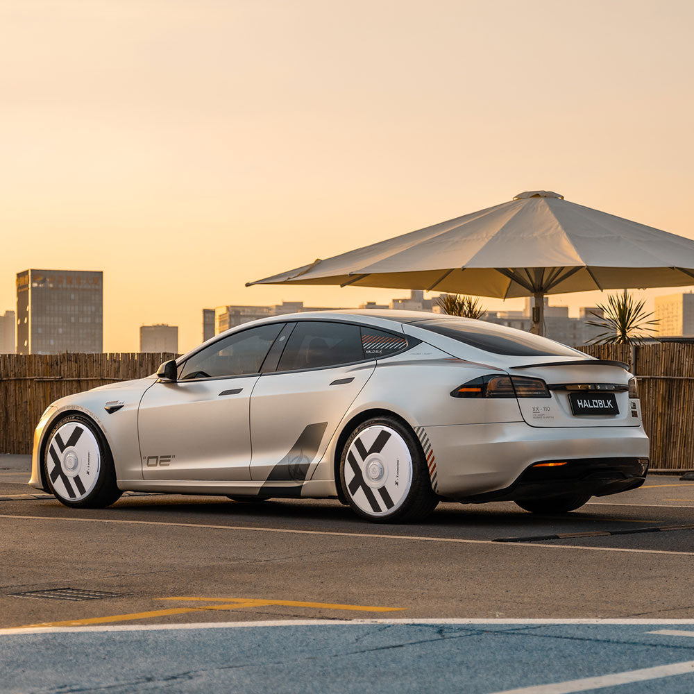 HALODISC 2 Wheel Covers for Tesla Model S 21" Arachnid Wheels by HALOBLK Store are shown on a sleek silver Tesla parked outdoors at sunset, with modern city buildings and a large umbrella in the background.