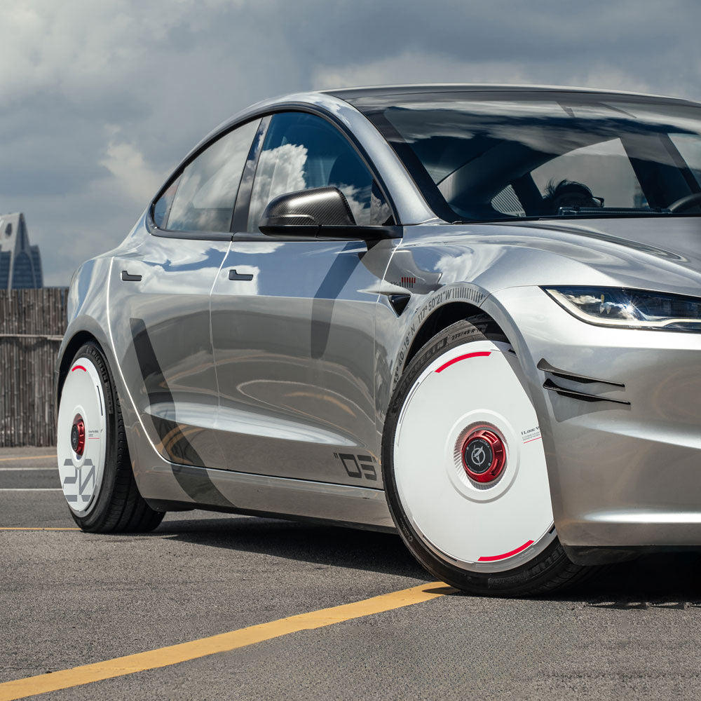 A sleek silver car with HALODISC 2 Wheel Covers for Tesla from HALOBLK Store, featuring custom white numbers, is parked on a street, its shiny surface reflecting the cloudy sky and modern buildings.