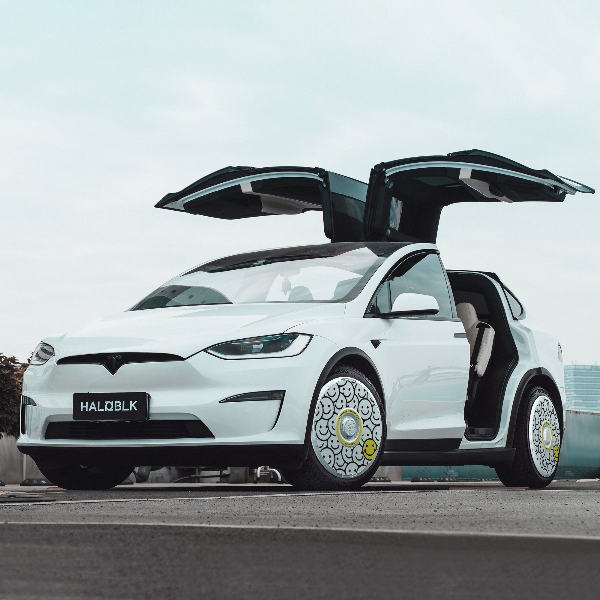 A white Tesla Model X (2021-2025) with its gull-wing doors open is parked on a rooftop, featuring HALODISC 2 "Lucky Day" wheel covers by HALOBLK Store on 20" Cyberstream wheels; a cityscape and overcast sky are seen in the background.