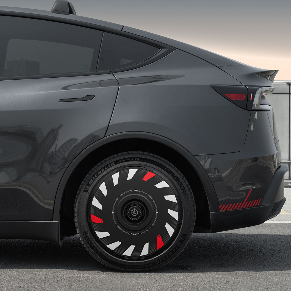A close-up of a dark gray Tesla Model Y shows its rear wheel with HALODISC 2 Wheel Covers by HALOBLK Store, made for 20" Helix 2.0 Juniper Turbo wheels. The unique black cover features white and red geometric accents; parked by a concrete barrier.