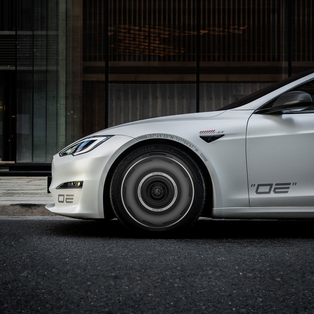 A white Tesla Model S with HALODISC 2 Wheel Covers (Vision-GT, Magnetite 19") from HALOBLK Store is parked on a city street, highlighting the front wheel and OE decals against a modern building backdrop.