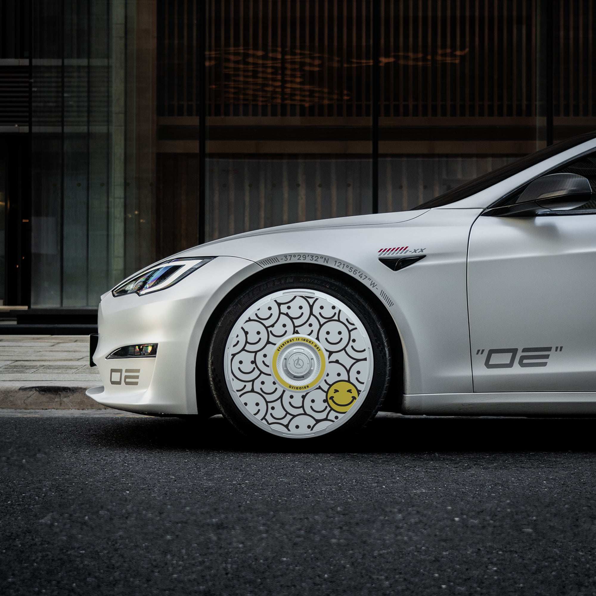 A white car with HALODISC 2 Wheel Covers for Tesla Model S 21" Arachnid Wheels by HALOBLK Store, featuring black smiley faces and a single yellow one near the bottom, is parked on the street against a modern building backdrop.