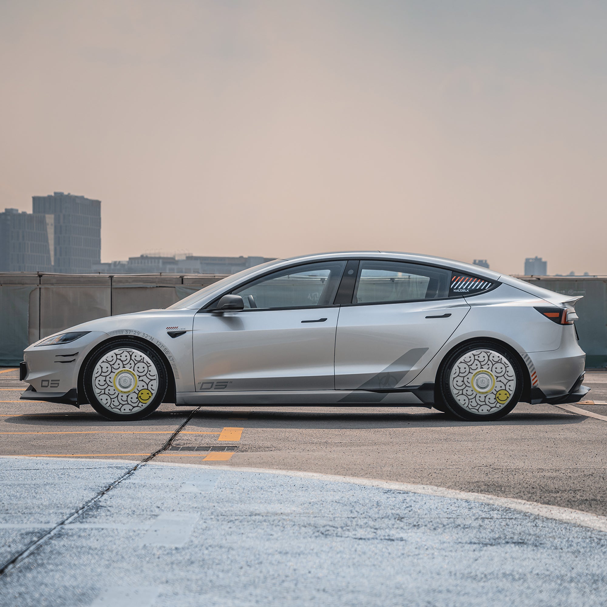 A silver Tesla Model 3 Highland with HALODISC 2 Wheel Covers by HALOBLK Store is parked on a rooftop, city buildings and a hazy sky in the background.