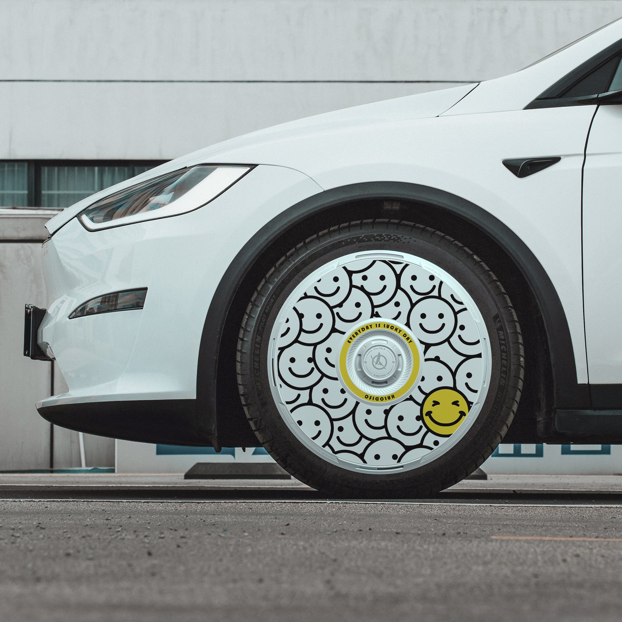A white Tesla Model X with HALOBLK Store's HALODISC 2 "Lucky Day" wheel covers on its 20" Cyberstream wheels—featuring a decorative design of black-outlined smiley faces with one yellow smiley face.