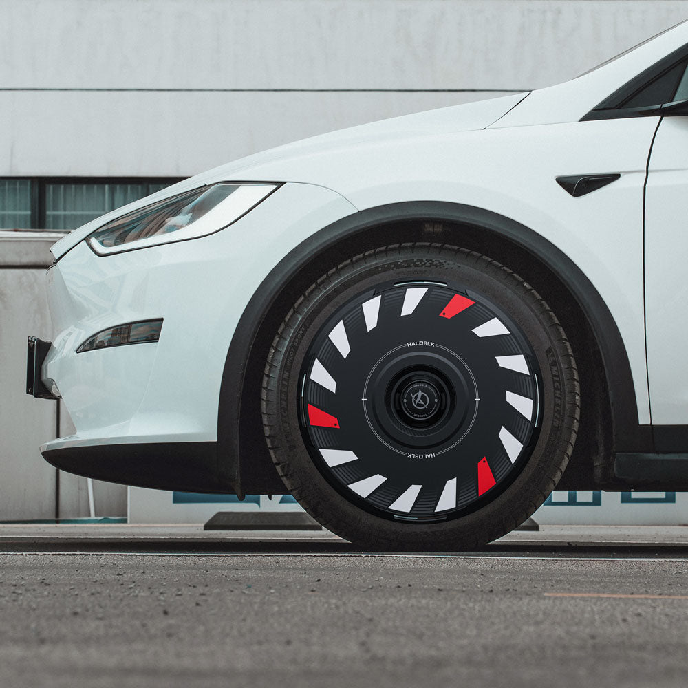 A close-up side view of a white Tesla Model X (2016-2020) with 20" Slipstream wheels featuring HALODISC 2 Turbo wheel covers from HALOBLK Store, showcasing a black, white, and red geometric aerodynamic design, parked on asphalt by a modern building.