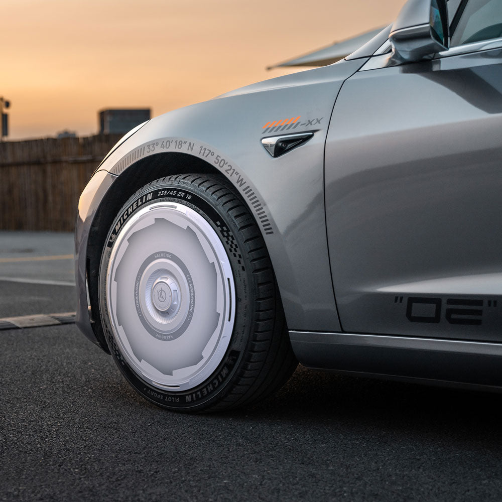 Close-up of HALODISC 2 Wheel Covers for Tesla Model 3 2018-2023 20" Performance Wheels by HALOBLK Store, featuring a futuristic white and gray rim design, installed on a silver car parked on asphalt at sunset with a wooden fence behind.