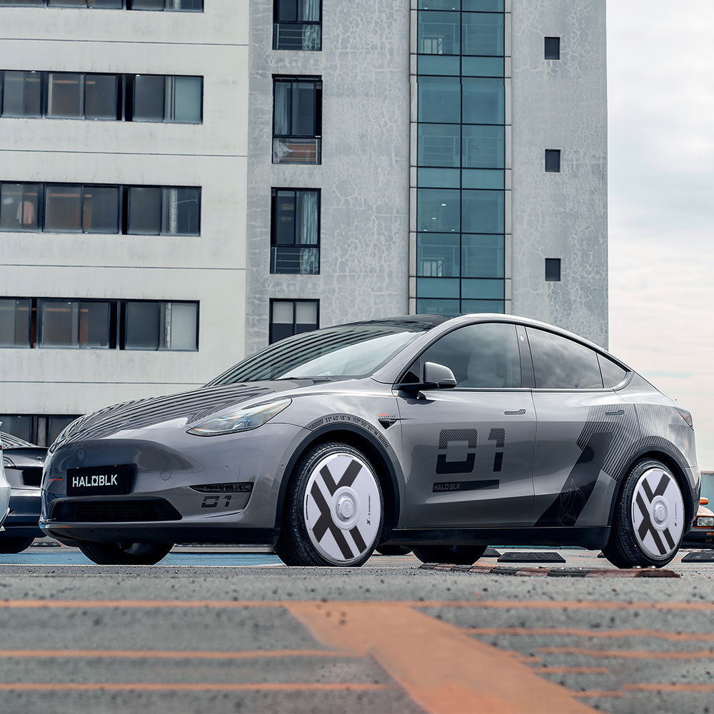 A Tesla Model Y with HALODISC 2 Wheel Covers by HALOBLK Store is parked in the city. The 21" X-Factor wheel covers feature black and white geometric designs, with "HALOBLK" on the front plate and a modern high-rise in the background.