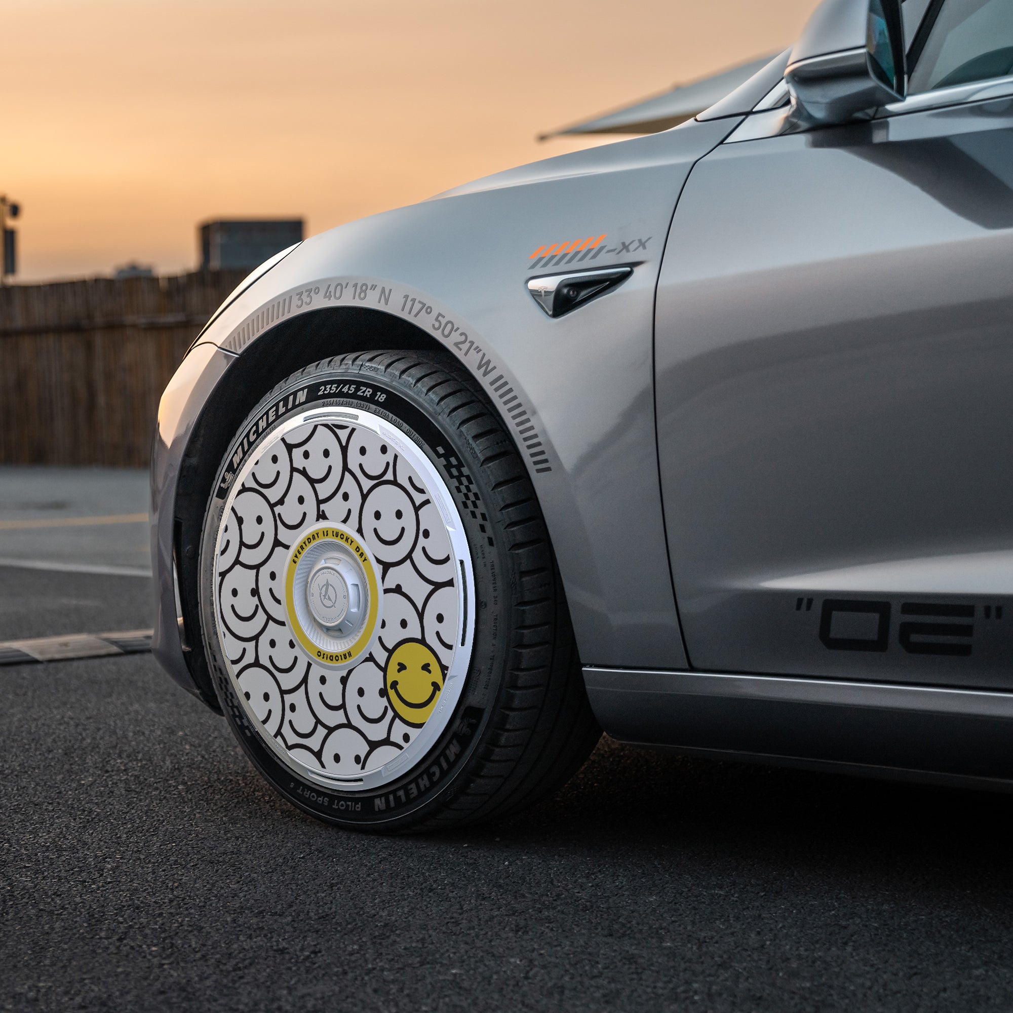 Close-up of a Tesla Model 3 (2019-2023) with HALODISC 2 “Lucky Day” 19" Zero-G wheel covers from HALOBLK Store, featuring black, white, and one yellow smiley face hubcap design, parked outdoors at sunset.