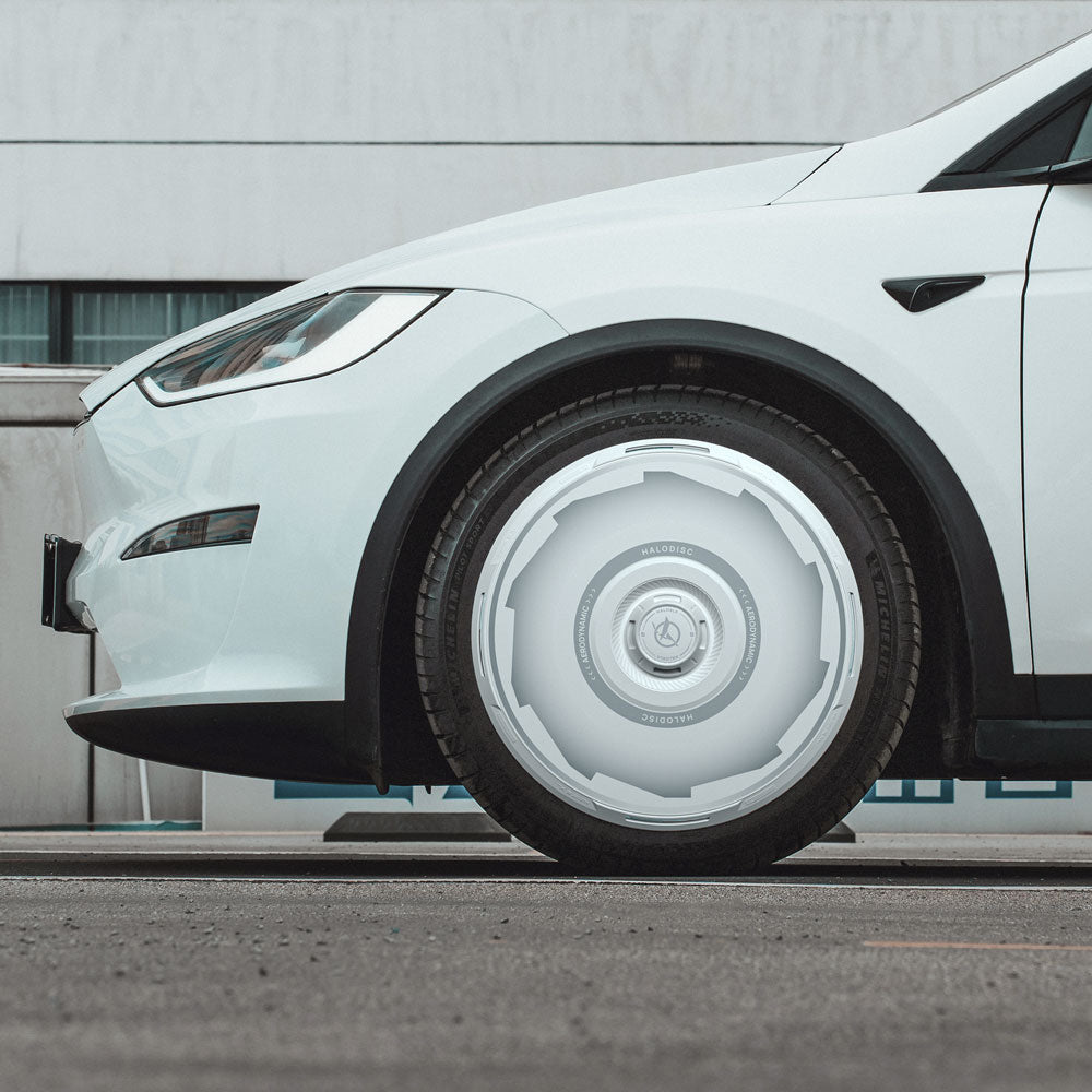 Close-up of a white Tesla Model X (2021-2025) with HALODISC 2 Vision-GT 20" wheel covers by HALOBLK Store, parked on asphalt. The front wheel shows the unique futuristic hubcap design; a white building with windows is in the background.