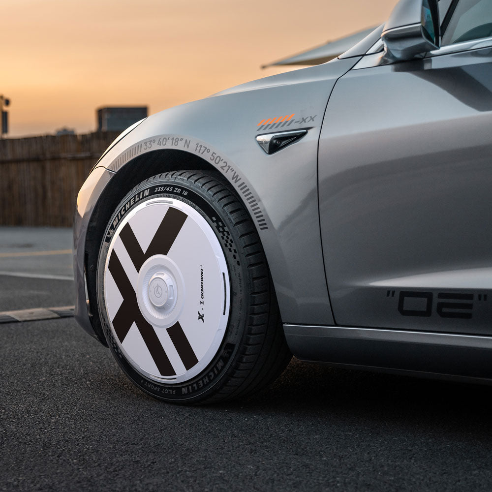 Close-up of a silver Tesla Model 3 (2017-2018) with HALODISC 2 X-Factor 19" wheel covers from HALOBLK Store, showcasing a bold black and white X-pattern and detailed tire branding at sunset.