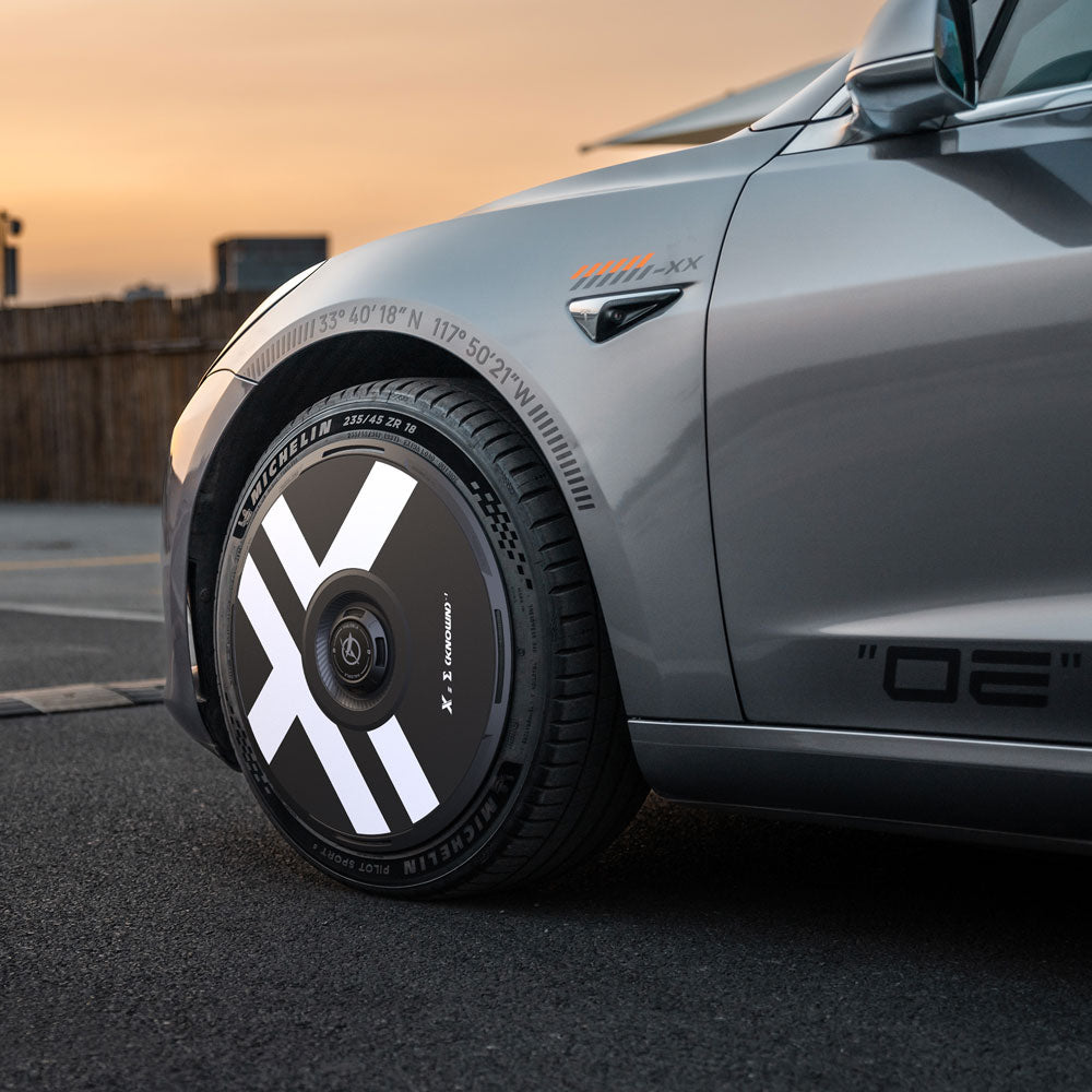 Close-up of HALODISC 2 Wheel Covers by HALOBLK Store with bold white X graphics on a Tesla Model 3 (2017-2018) 19" Performance wheel, parked at sunset near a wooden fence.