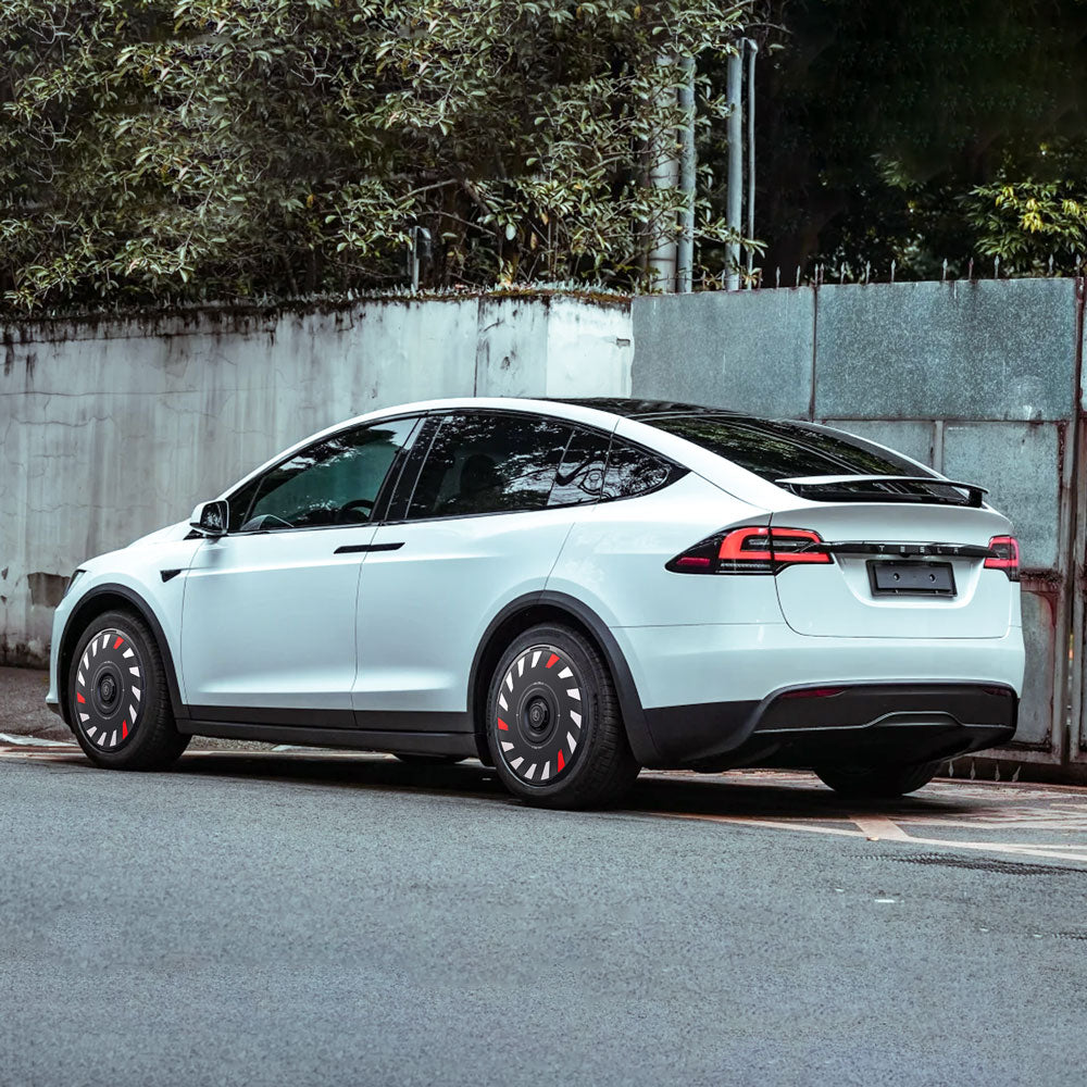 A white Tesla Model X with HALODISC 2 Wheel Covers by HALOBLK Store on 20" Slipstream wheels is parked by a concrete wall and metal gate, leafy trees behind it. The covers have black and red accents for a modern look.