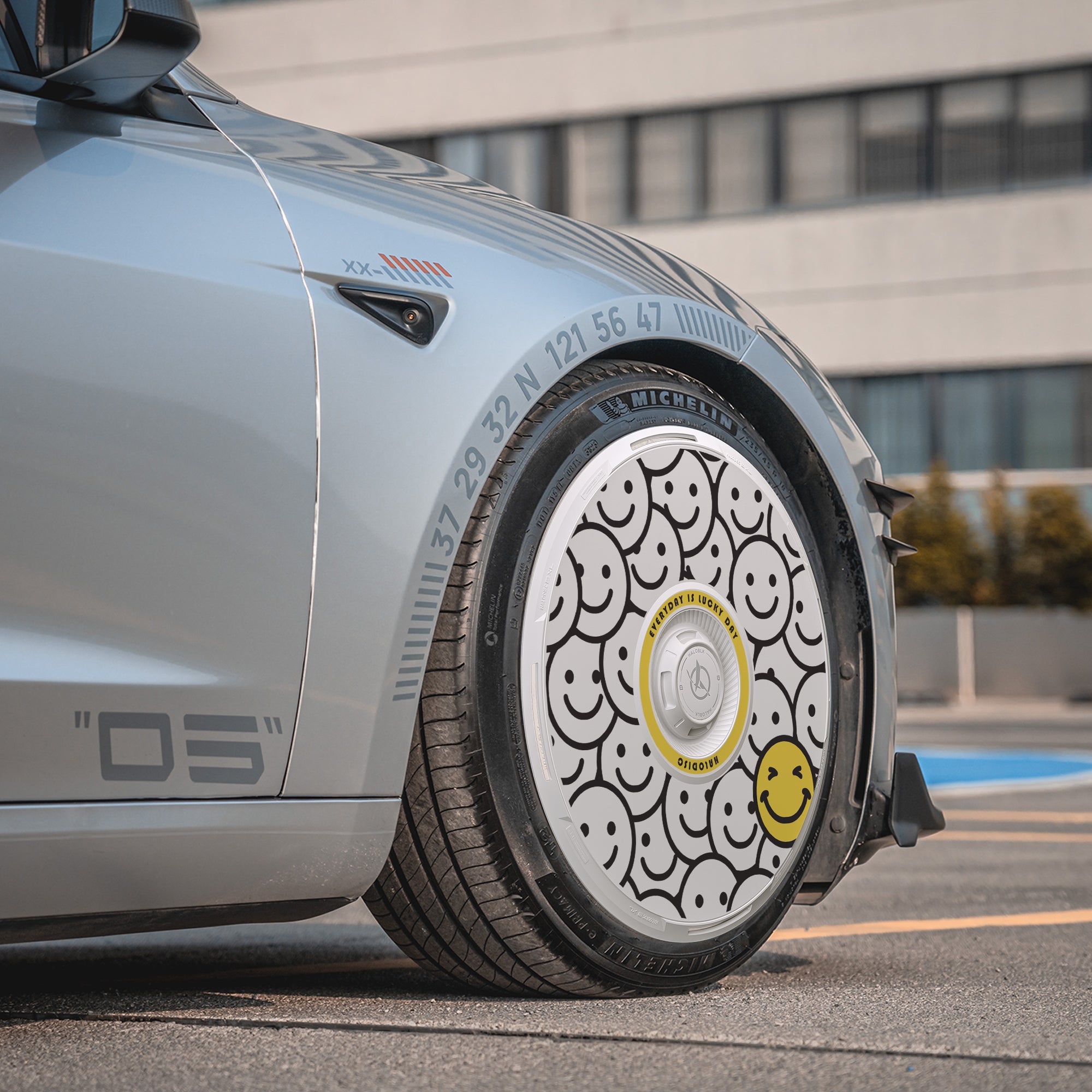 A close-up of a Tesla Model 3 Highland with HALODISC 2 Wheel Covers for 19" Nova Wheels by HALOBLK Store, featuring custom black outlined smiley faces and one yellow smiley face, parked on a city street.