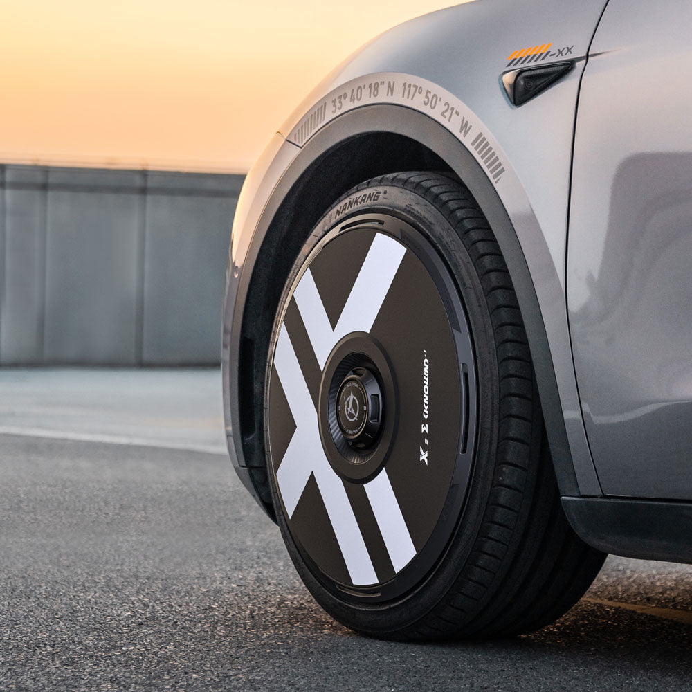 Close-up of HALODISC 2 Wheel Covers for Tesla Model Y 21" Überturbine Wheels by HALOBLK Store, featuring black and white X-Factor aerodynamic rims with bold X-shaped white stripes, on a silver car with graphic accents at sunset.