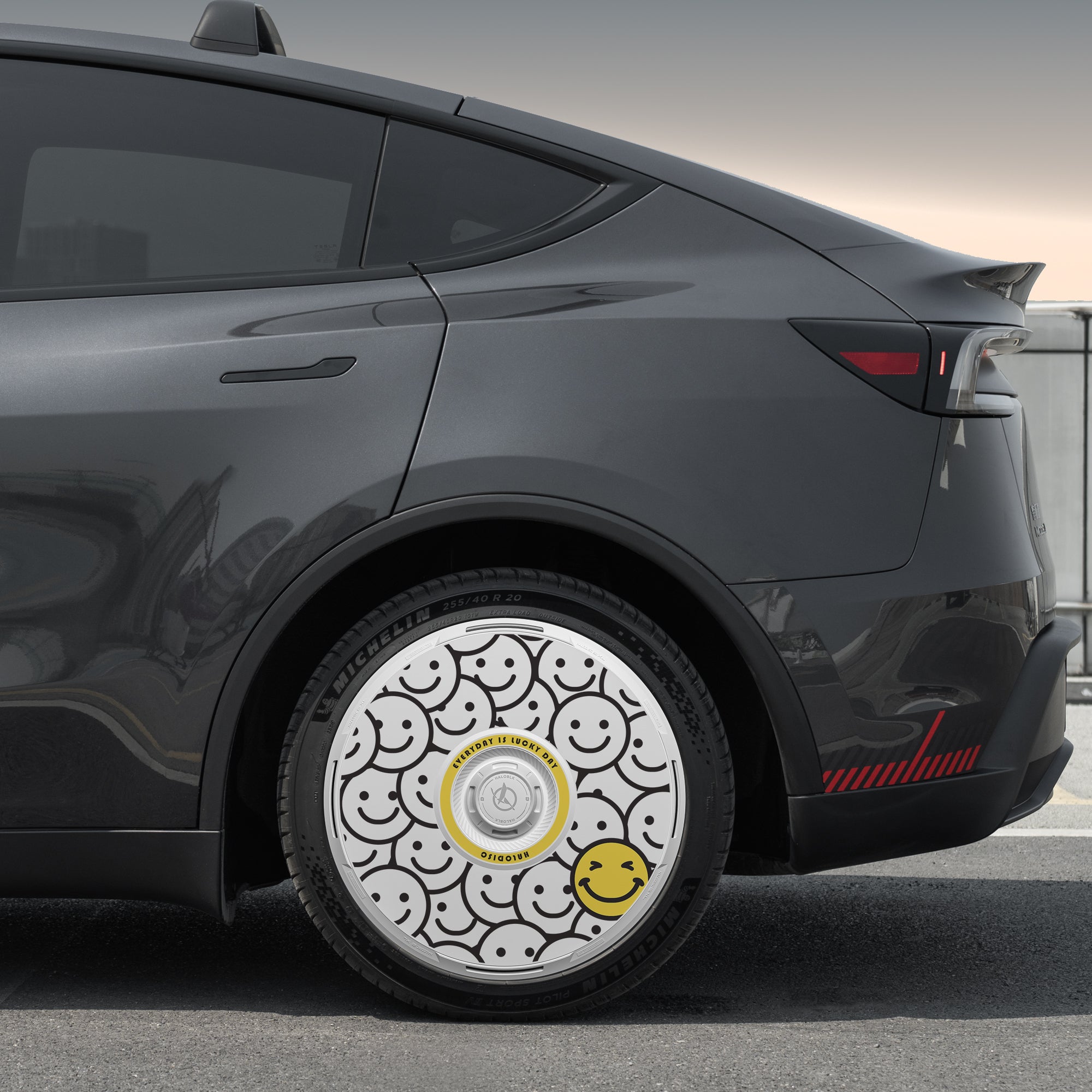 A close-up of a Tesla Model Y Juniper 19" Crossflow wheel with the HALODISC 2 "Lucky Day" wheel cover from HALOBLK Store, featuring black smiley faces on white and a standout large yellow smiley face.