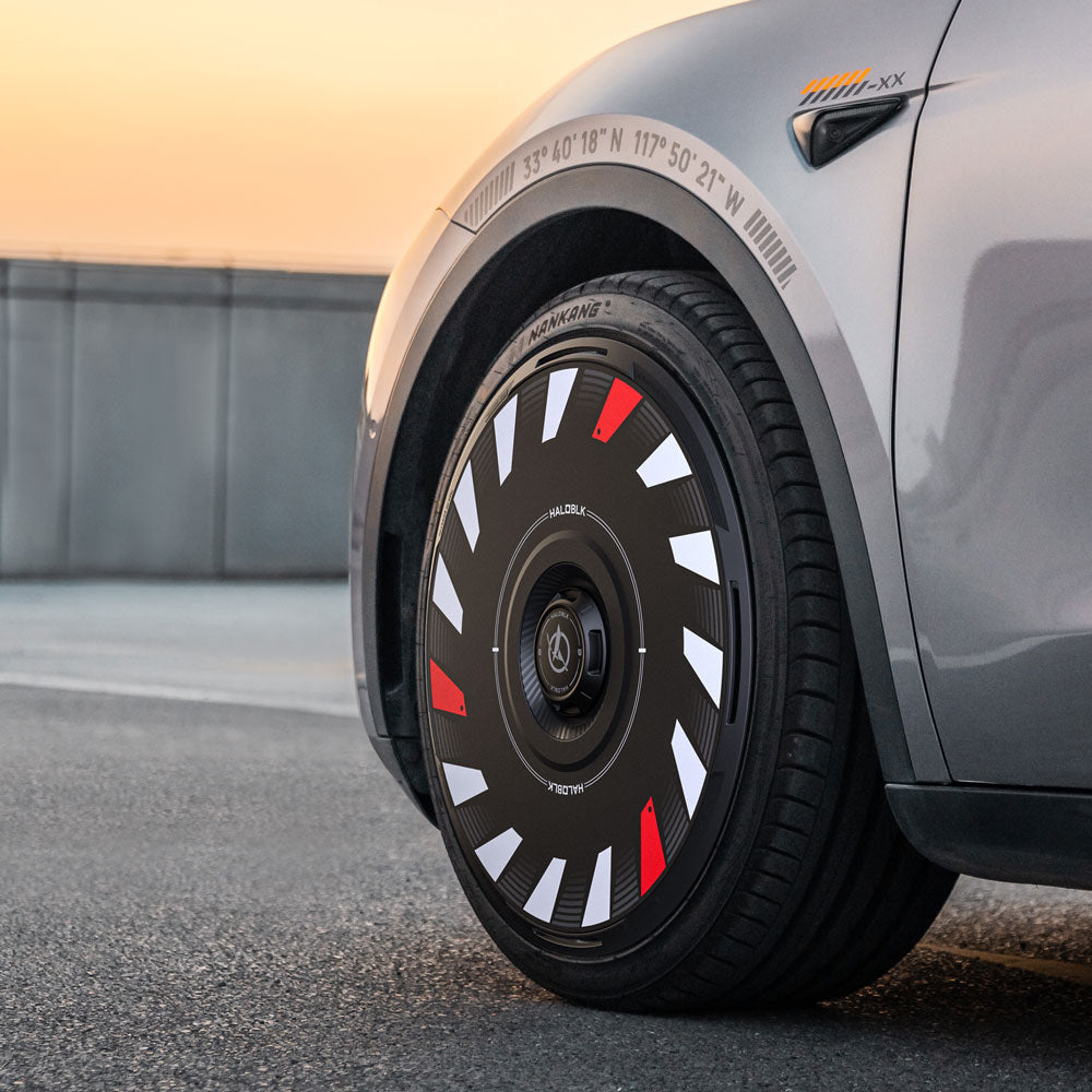 Close-up of HALODISC 2 Wheel Covers by HALOBLK Store on a Tesla Model Y 20" Induction Wheel. The unique black cover with white and red accents stands out beneath bold coordinates on the silver fender, captured outdoors at sunset.