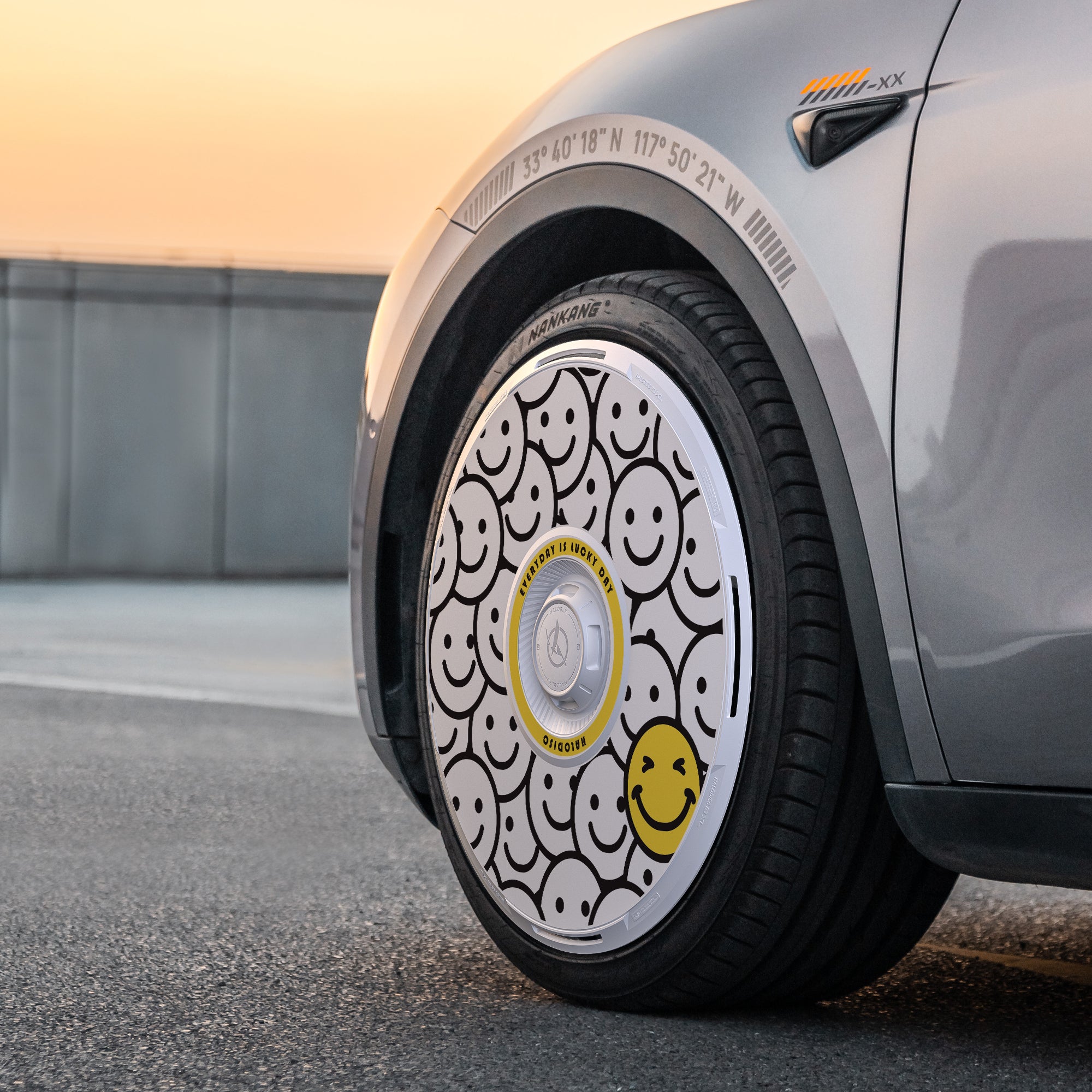 A close-up of the HALODISC 2 Wheel Covers for Tesla Model Y 21" Überturbine Wheels (Lucky Day) by HALOBLK Store, featuring a white hubcap with multiple black and one large yellow smiley face, parked on a road at sunset.