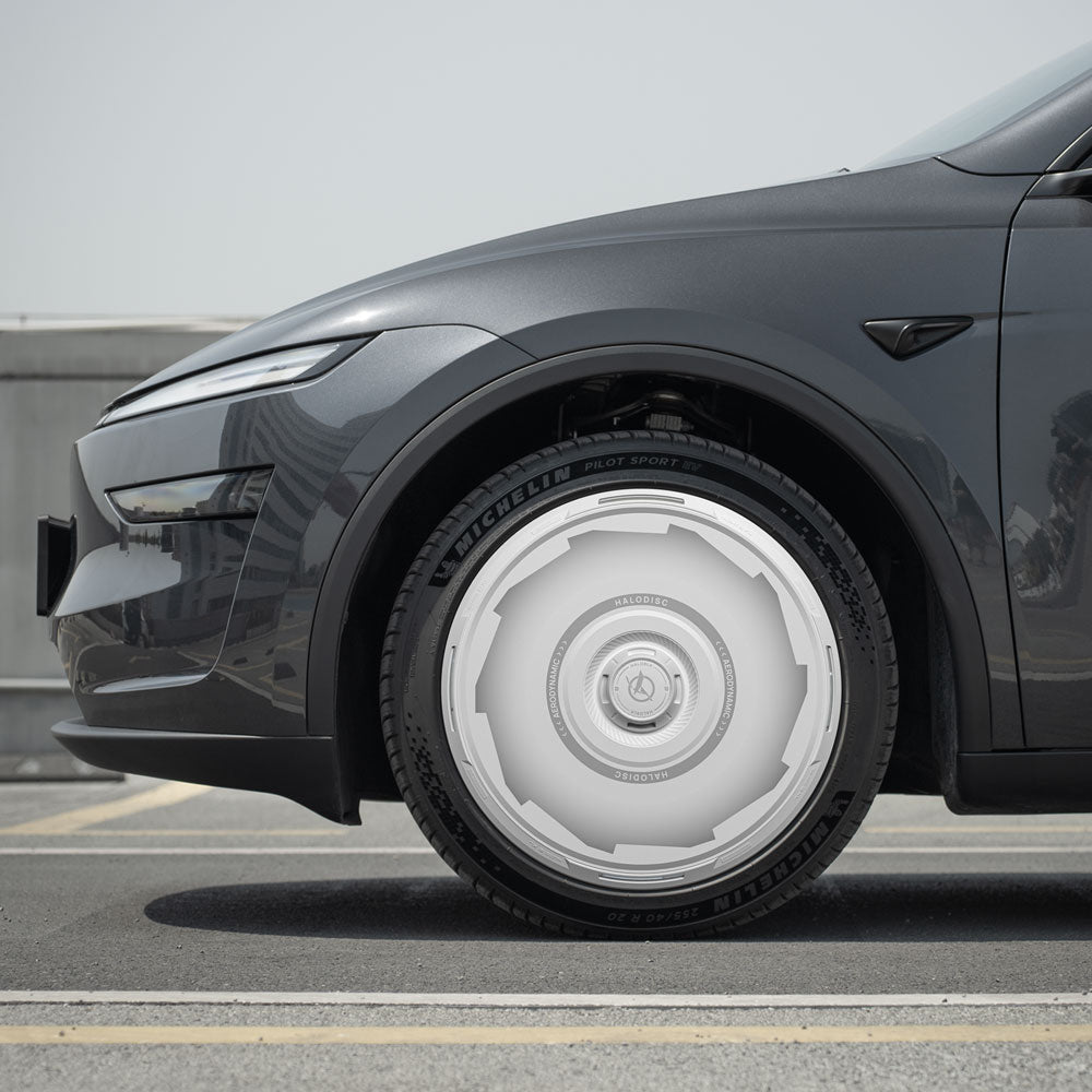 A close-up of the front left wheel of a dark gray Tesla Model Y with HALODISC 2 Vision-GT 19" Crossflow silver wheel covers by HALOBLK Store, parked on a rooftop under a clear sky.