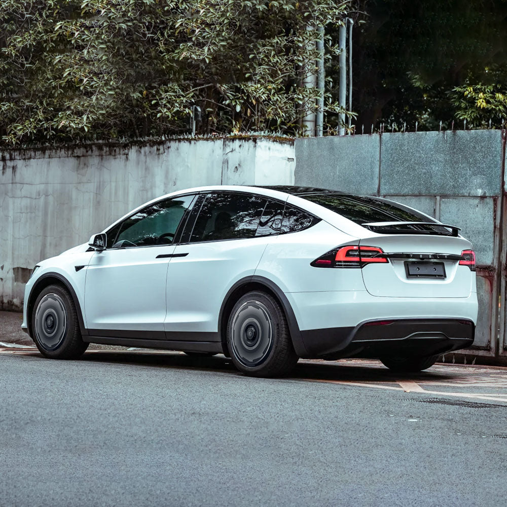 A white Tesla Model X (2021-2025) with HALODISC 2 Wheel Covers for 20" Cyberstream Wheels by HALOBLK Store is parked on a street beside a tall white wall and trees, with no visible license plate.