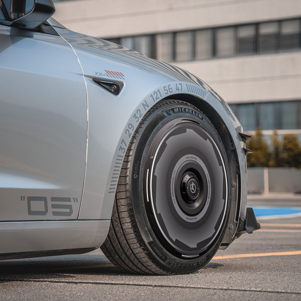 A close-up of HALODISC 2 Wheel Covers for Tesla Model 3 Highland 20" Warp Wheels by HALOBLK Store, showcasing their sleek, modern design with circular markings, installed on a parked car near a modern building.
