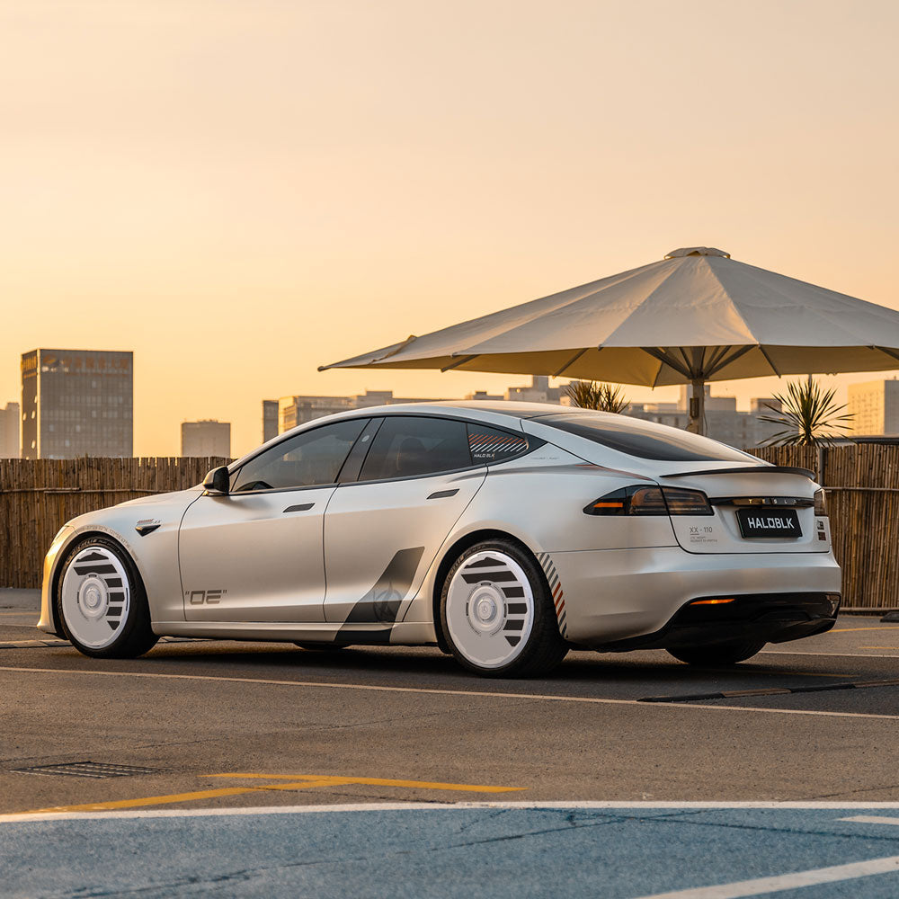 A Tesla Model S with HALODISC 2 Wheel Covers (19" Slipstream, HALOBLK Store) is parked outdoors at sunset near a white umbrella and fencing, with city buildings in the background; license plate reads HALBO1K.