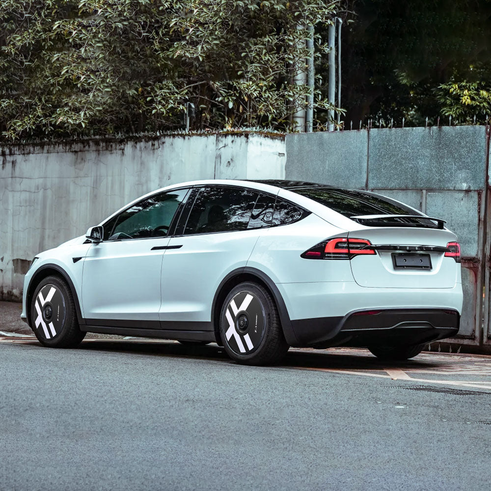 A white Tesla Model X (2021-2025) with HALODISC 2 Wheel Covers by HALOBLK Store on 20" Cyberstream Wheels is parked near a concrete wall and greenery, viewed from the rear side angle.