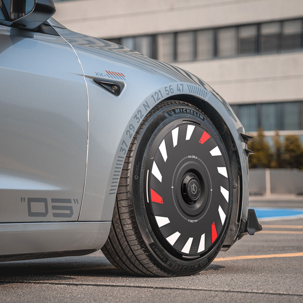 A close-up of HALODISC 2 Wheel Covers by HALOBLK Store for Tesla Model 3 Highland 18" Photon Wheels, showcasing the unique design with white, red, and black accents, parked on a street with a modern building in the background.