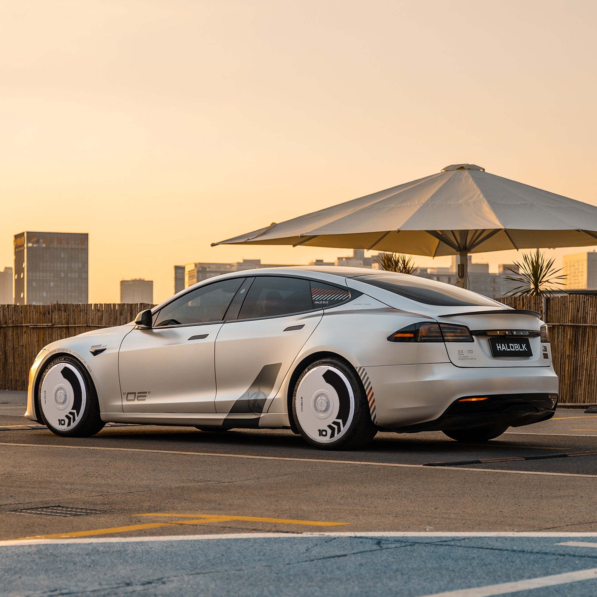 A silver Tesla Model S with HALODISC 2 Arrow white wheel covers by HALOBLK Store is parked in an urban lot at sunset, city buildings and a large outdoor umbrella in the background.