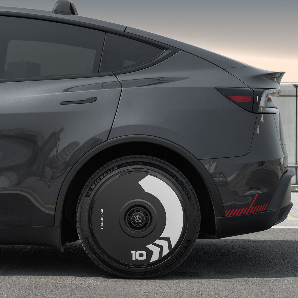A close-up side view of a dark gray car parked on a rooftop showcases HALODISC 2 Wheel Covers for Tesla Model Y Juniper 20" Helix 2.0 Wheels by HALOBLK Store, featuring bold black and white geometric Arrow patterns with the number “10” on the rear wheel.