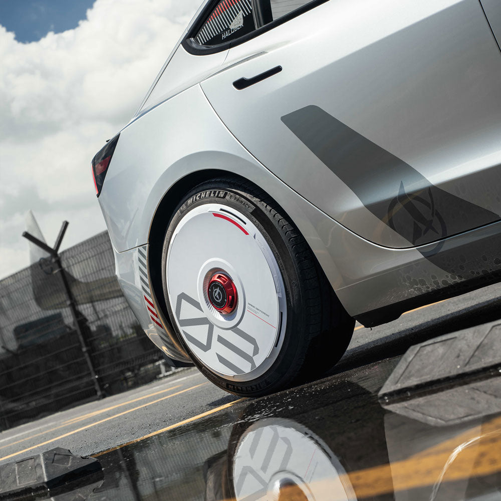 A close-up of a Tesla’s rear wheel with HALODISC 2 Wheel Covers in white from HALOBLK Store, featuring custom numbers, parked near a reflective puddle; an airport runway and cloudy sky appear in the background.