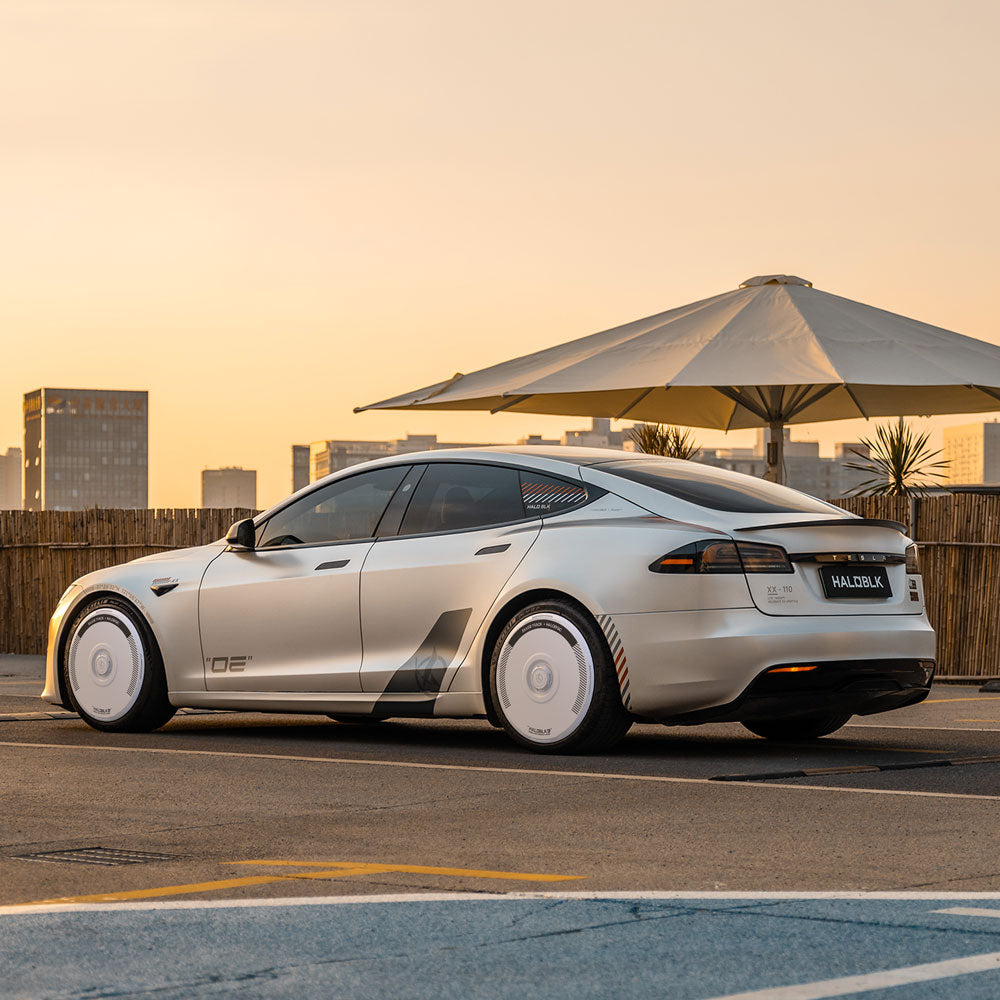 A Tesla Model S with HALODISC 2 Silver Track Wheel Covers by HALOBLK Store is parked in an outdoor lot at sunset, city buildings and a large umbrella in the background highlight its stylish, futuristic design.