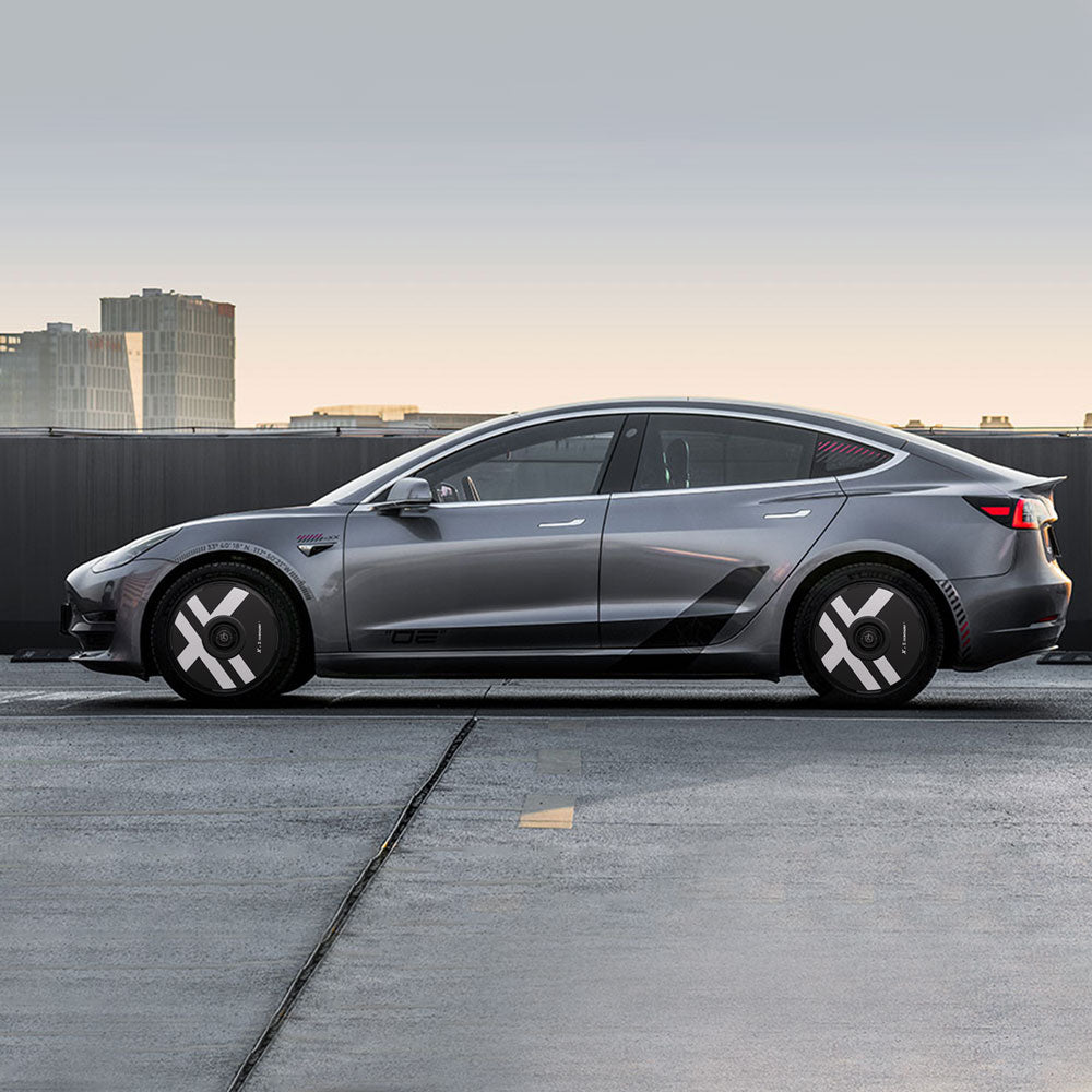 A silver Tesla Model 3 (2018-2023) with HALODISC 2 X-Factor wheel covers by HALOBLK Store, featuring geometric black and white designs on the wheels, is parked on a rooftop against a city skyline at sunset.