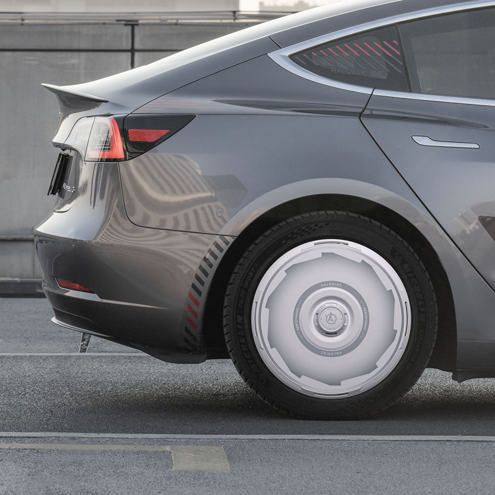 A close-up of a gray Tesla Model 3 (2019-2023) parked on the street highlights the futuristic silver HALODISC 2 Wheel Covers by HALOBLK Store, designed for 19" Zero-G Wheels. In the background are a concrete barrier and part of a building.