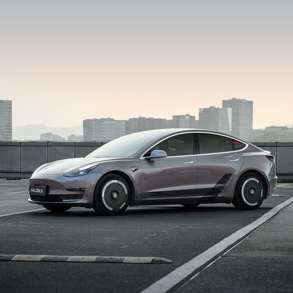 A silver Tesla Model 3 (2017-2018) with HALODISC 2 Wheel Covers by HALOBLK Store in Silver Track is parked on a rooftop at dusk, city skyline behind, featuring aerodynamic hubcaps and a sleek reflective finish.