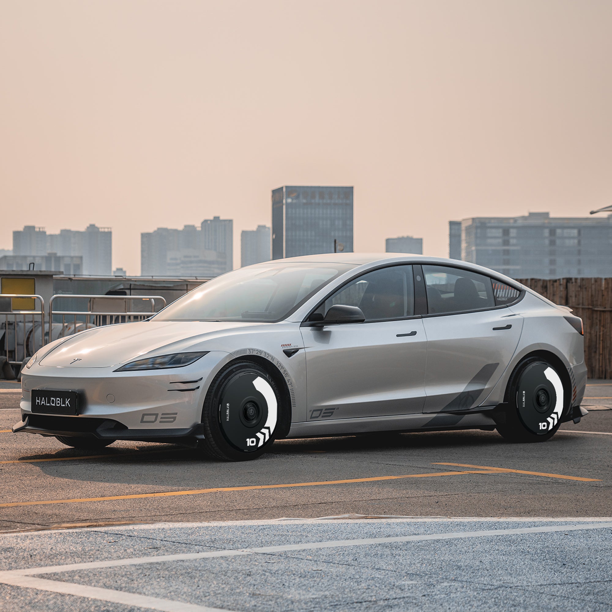 A Tesla Model 3 Highland with HALODISC 2 Arrow wheel covers from HALOBLK Store is parked on an urban rooftop lot, showcasing its stylish black and white 18" Photon wheels against a backdrop of tall modern buildings and a hazy sky.