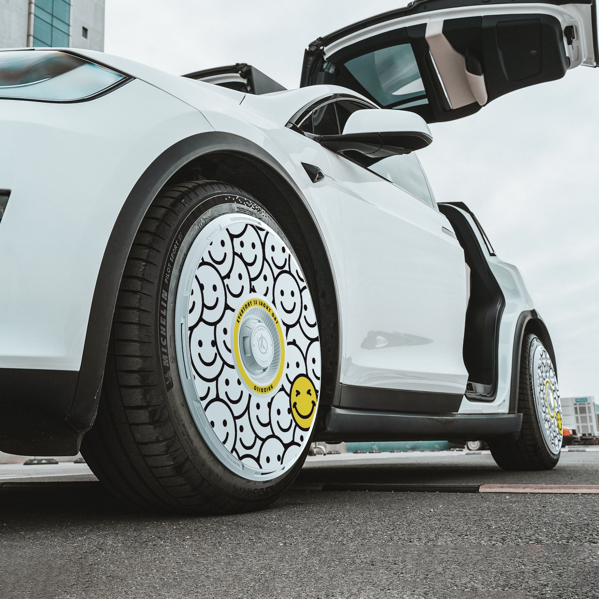 A white Tesla Model X (2021-2025) with an open gull-wing door sports HALODISC 2 "Lucky Day" wheel covers from HALOBLK Store, featuring black and white smiley face patterns and a large yellow smiley, shot from a low angle.